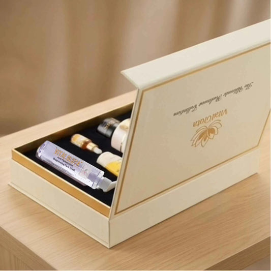 An open vitalgluta cream-colored gift box with gold trim on a light wood table, containing several small cosmetic or essential oil vials. The inside lid is printed with elegant gold script and a floral emblem.