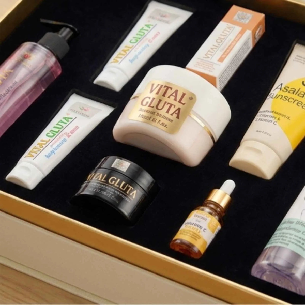 A skincare gift set arranged in a black-lined box with a gold border. The set includes a pink bottle, white and orange VITAL GLUTA tubes and box, a white VITAL GLUTA cream jar, a black VITAL GLUTA jar, a small amber dropper bottle, and an Asala sunscreen tube.