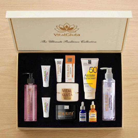 VitalGluta skincare collection gift set in an open cream-colored box with a black interior, containing multiple skincare products including a pink bottle, white tubes, a yellow sunscreen tube, and small dropper bottles on a light wood background.
