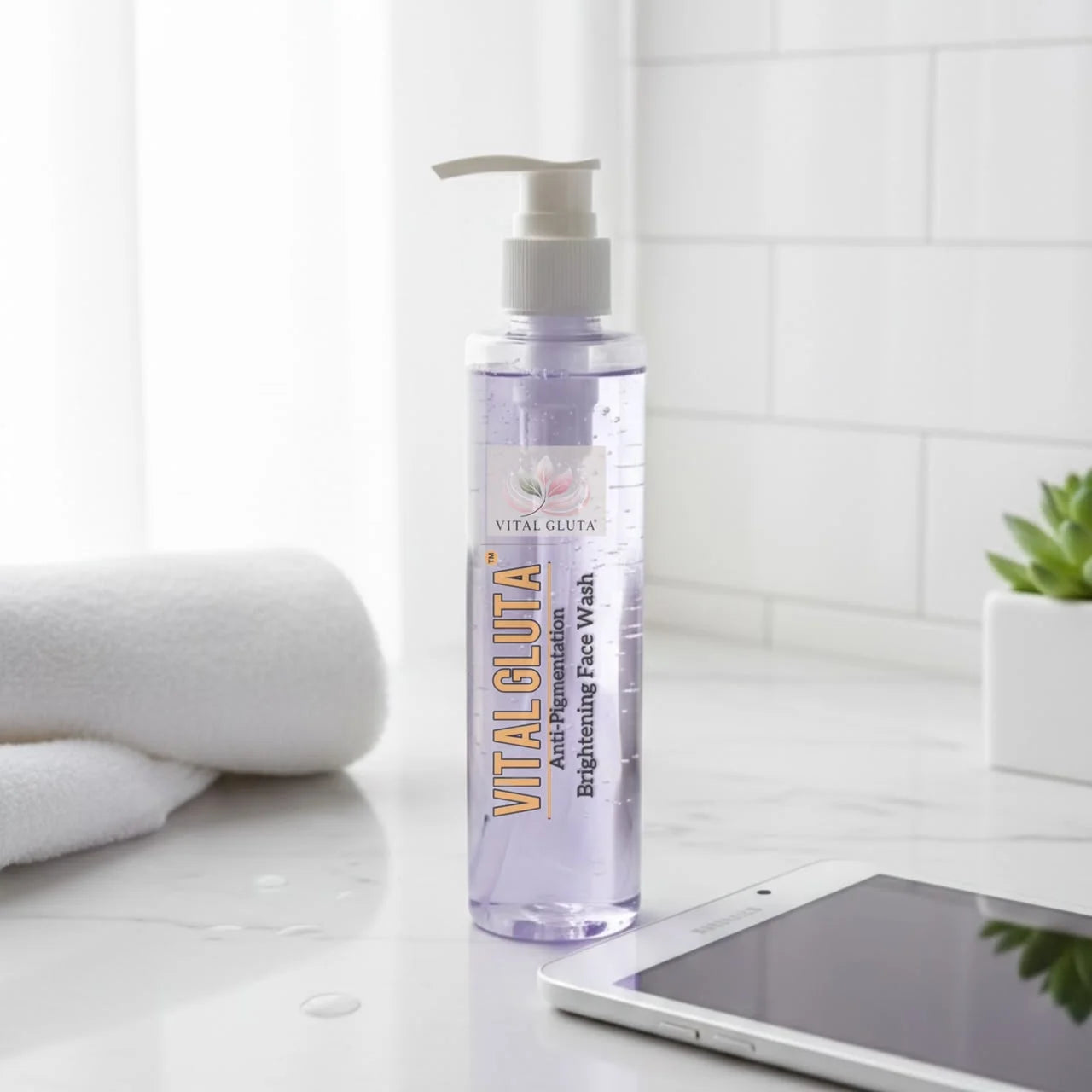 A bottle of Vital Gluta Brightening Face Wash standing on a white countertop in a bright bathroom-like setting, with a folded towel, tiled wall, small plant, and part of a tablet visible.