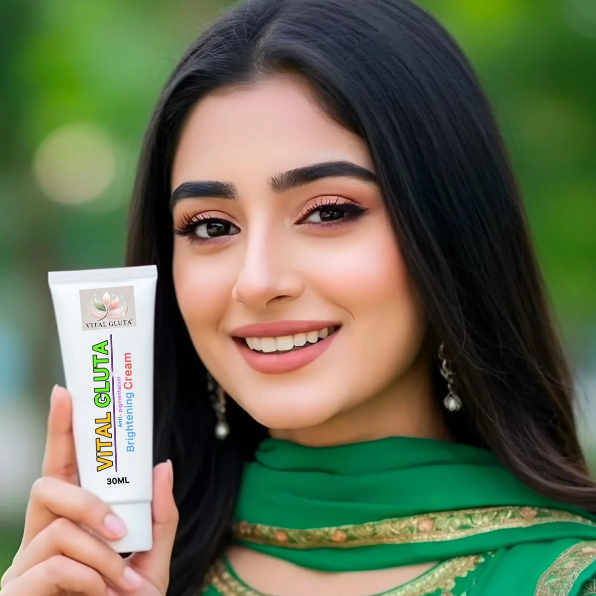 A smiling woman with long black hair holds up a white tube of Vital Gluta Brightening Cream. She is wearing a green traditional outfit with gold embroidery, set against a soft, blurred green background.